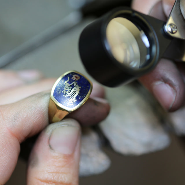A jeweller looking at a bespoke yellow gold signet ring with blue enamel and a dragon design through a magnifying glass.